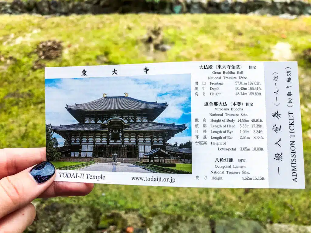 A hand holds an admission ticket to Todai-ji Temple in Nara, featuring an image of the Great Buddha Hall and detailed measurements of the statues and building.