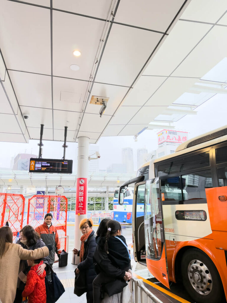 Passengers line up beside an orange and white airport limousine bus at a covered outdoor terminal in Japan. A digital sign overhead shows bus stop A1 at 13 50 while travelers with coats and luggage prepare to board. This is the airport limousine bus from Shinjuku station to Haneda airport.
