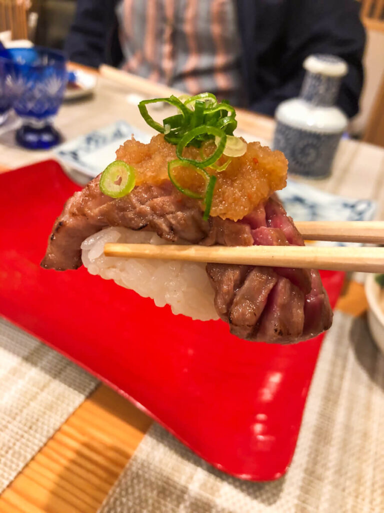 Chopsticks holding a piece of seared beef nigiri topped with grated ginger and sliced green onions over sushi rice. The close up highlights the tender marbled meat and glossy topping at a Japanese sushi restaurant.