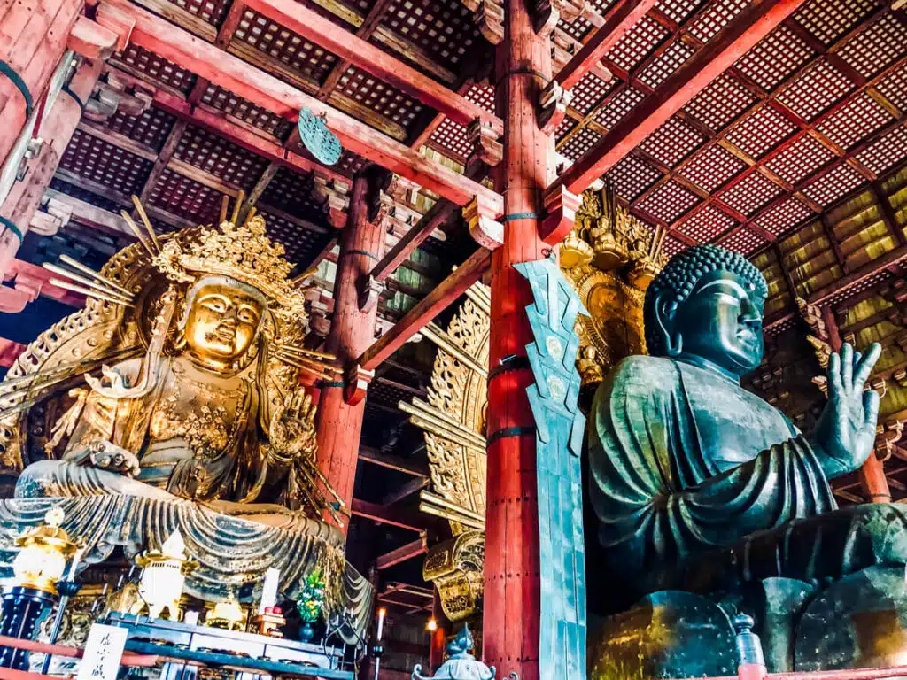 Ornate golden and bronze Buddha statues inside Todai-ji Temple in Nara, Japan, framed by tall red wooden beams and a detailed coffered ceiling.