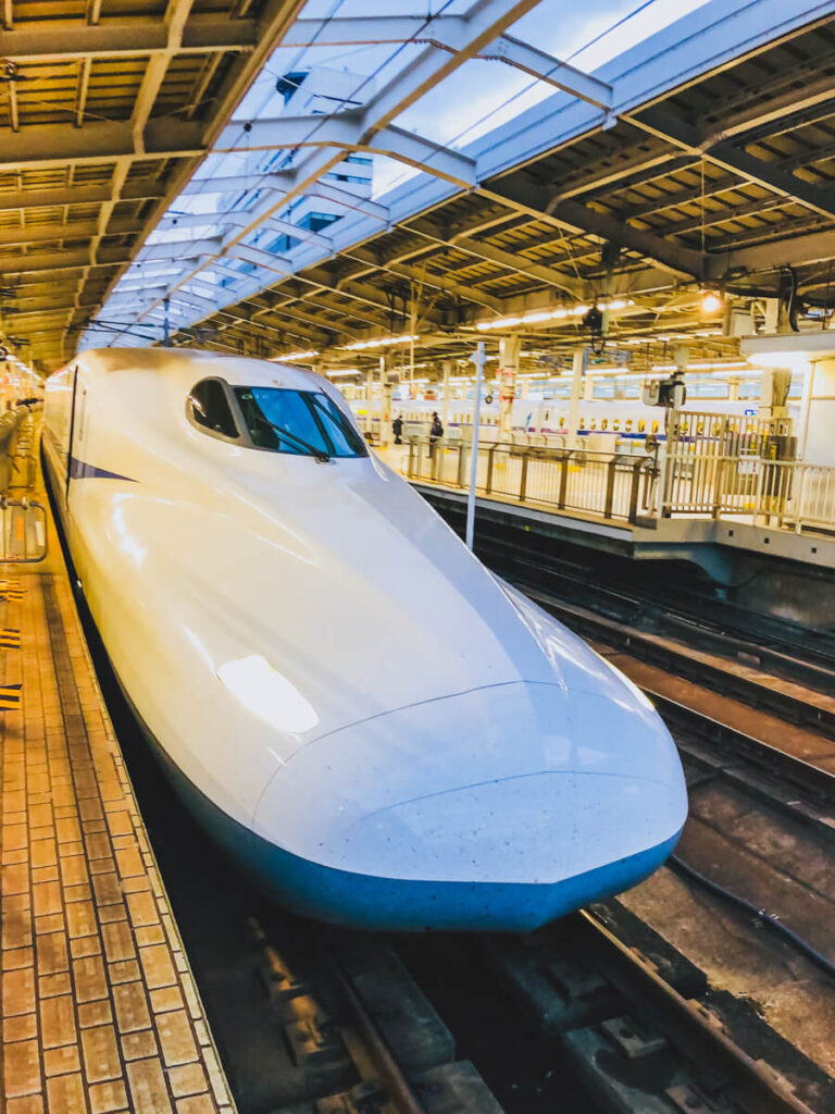 White bullet train parked at a station platform under bright overhead lights with its sleek pointed nose facing forward. The modern high speed train sits on the tracks beside a tiled platform, highlighting Japan rail travel.