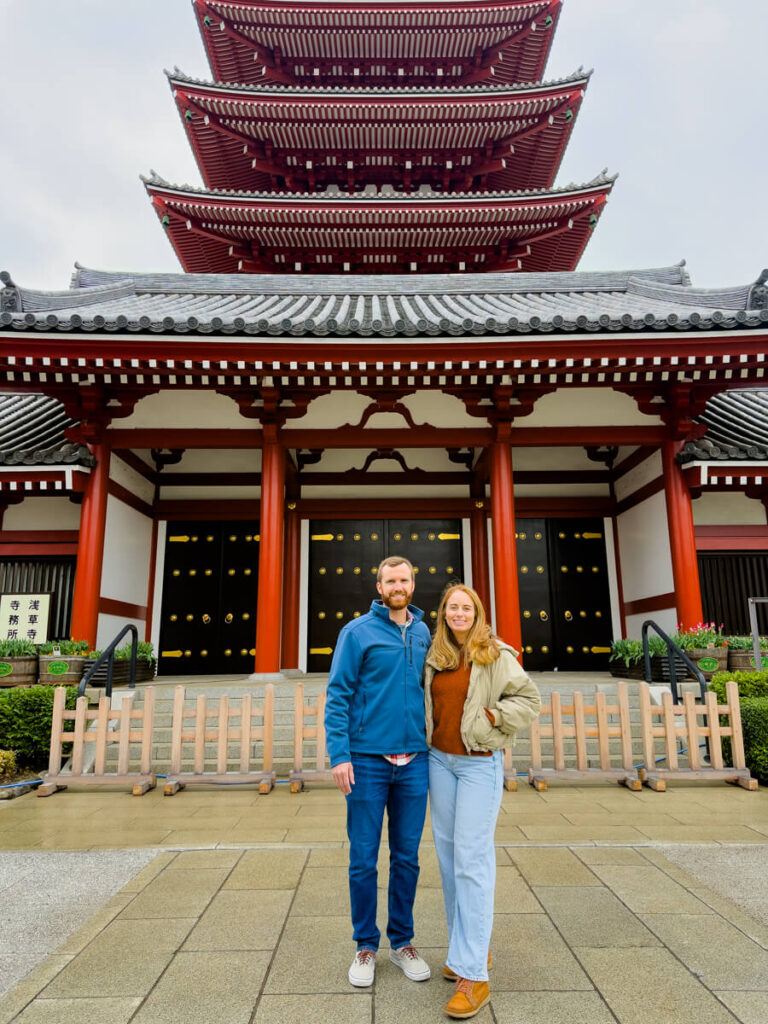 Couple standing in front of a red five story pagoda at Senso-ji temple with black and gold doors and traditional tiled roof. The iconic Japanese temple architecture towers above them in a spacious courtyard.