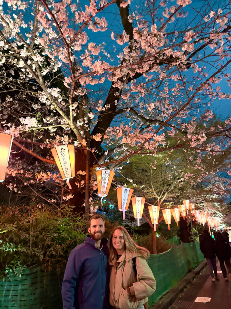A couple poses under rows of illuminated lanterns and cherry blossom trees in Ueno Park, enjoying a magical evening walk during sakura season in Japan.