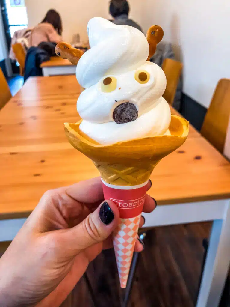 Soft serve ice cream in a cone decorated to resemble a surprised deer face, with cookie antlers, chocolate candy eyes, and a round chocolate nose, held in a cozy cafe setting in Nara. The design playfully references the famous Nara deer