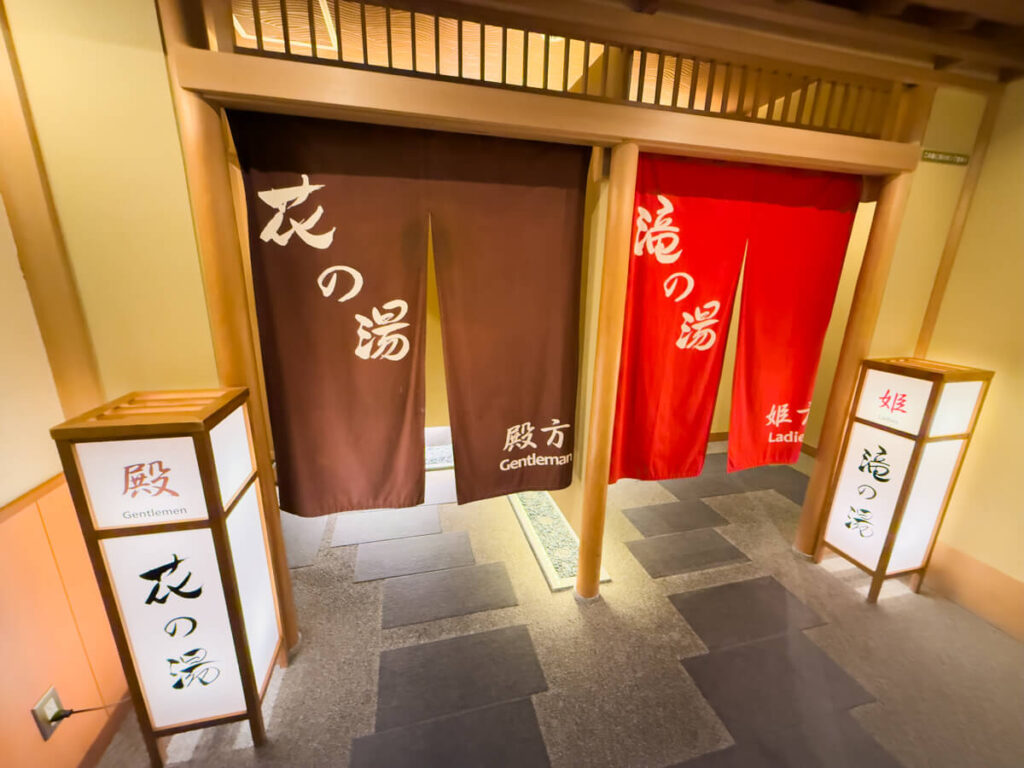 Entrance to a ryokan onsen area with fabric noren curtains—brown labeled “Gentlemen” and red labeled “Ladies”—hanging over separate doorways. Soft lighting and wooden accents create a calm atmosphere.