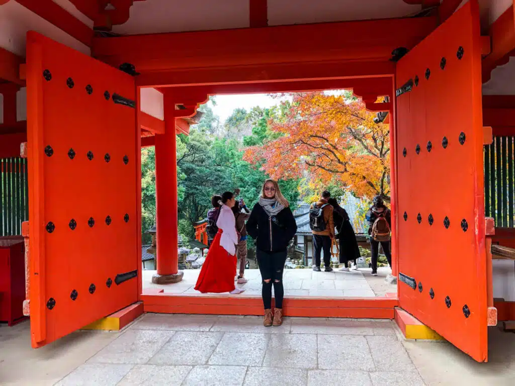 A woman stands between the large open red doors of Kasugataisha Shrine, framed by autumn foliage and visitors in traditional and modern clothing.