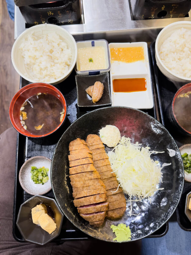 Japanese set meal with sliced breaded beef cutlet served with shredded cabbage rice miso soup and dipping sauces. The tray includes small side dishes and condiments arranged neatly around the main plate.