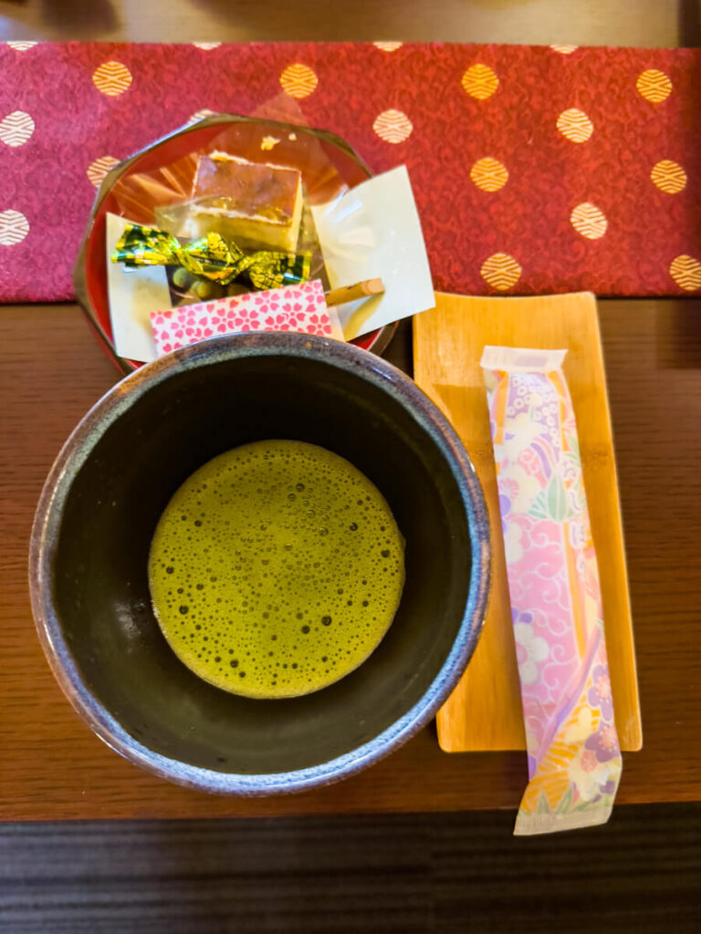 Ceramic bowl of frothy green matcha served with traditional Japanese sweets and a wooden pick on a tray. The vibrant tea and delicate confections create a classic tea ceremony style presentation.