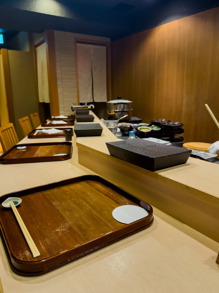 Minimalist Japanese sushi counter with light wood paneling and a row of wooden trays set with chopsticks and small dishes, ready for guests. The intimate restaurant setting shows a clean prep area behind the counter with neatly arranged cookware and ingredients, highlighting a traditional omakase dining experience.
