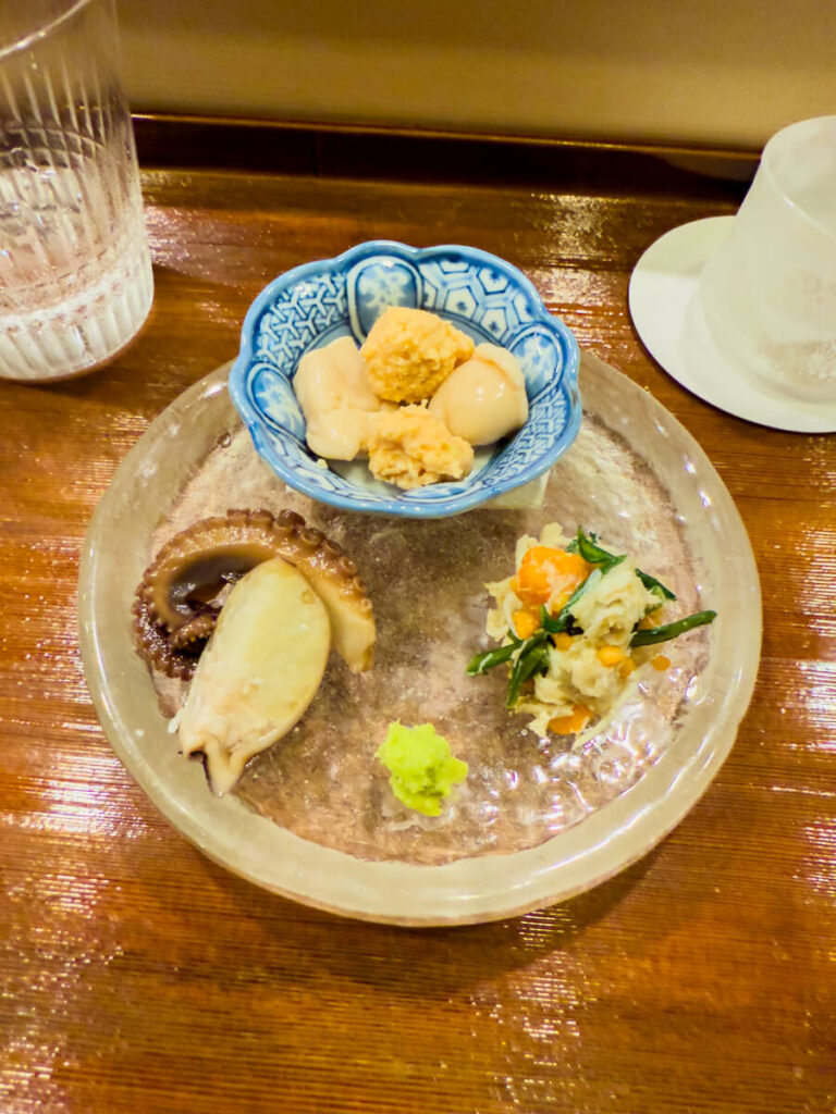 Assorted Japanese appetizers served on a clear glass plate at a wooden counter, featuring octopus tentacle, sliced seafood, a small mound of wasabi, and a mixed vegetable side. A blue patterned bowl holds simmered items like milt, capturing the delicate presentation of a multi course Japanese meal.