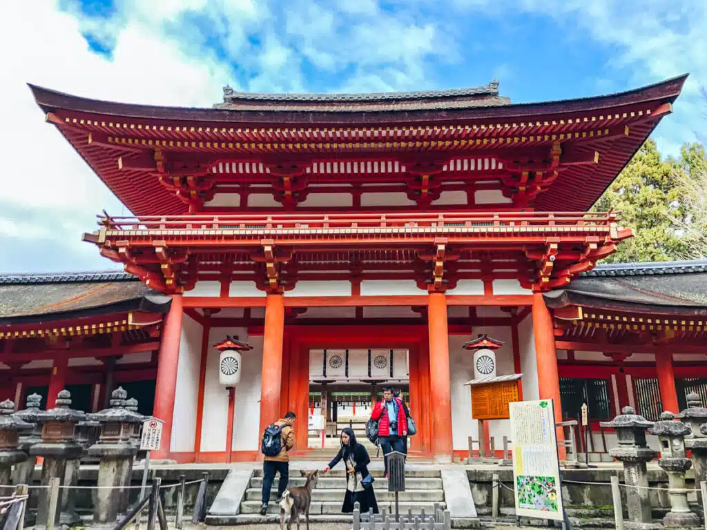 Bright red and white entrance gate of Kasugataisha Shrine in Nara, Japan, with a few visitors and a Nara deer near the stone steps.