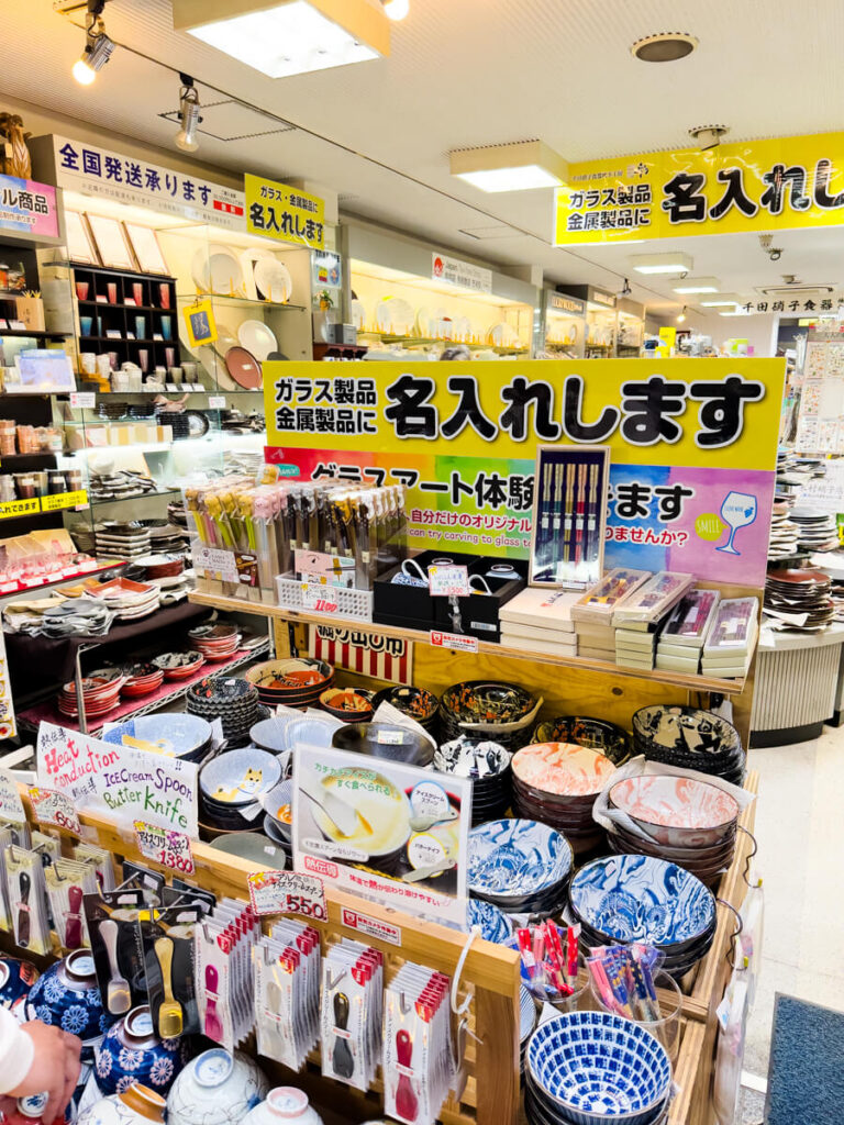 Colorful Japanese kitchenware and ceramic bowls displayed in a busy shop along Sennichimae Doguyasuji in Osaka, with shelves packed tightly with plates, chopsticks, and specialty utensils for restaurants. Bright yellow signs in Japanese advertise engraving services for glass and metal products, highlighting the customization options available in this famous kitchenware street.