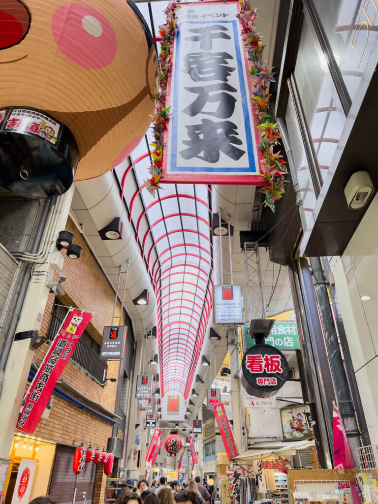 Covered shopping arcade at Sennichimae Doguyasuji in Osaka lined with hanging signs, red lanterns, and vertical banners written in Japanese. The long arched ceiling stretches above the bustling pedestrian walkway, capturing the lively atmosphere of this well known restaurant supply street.