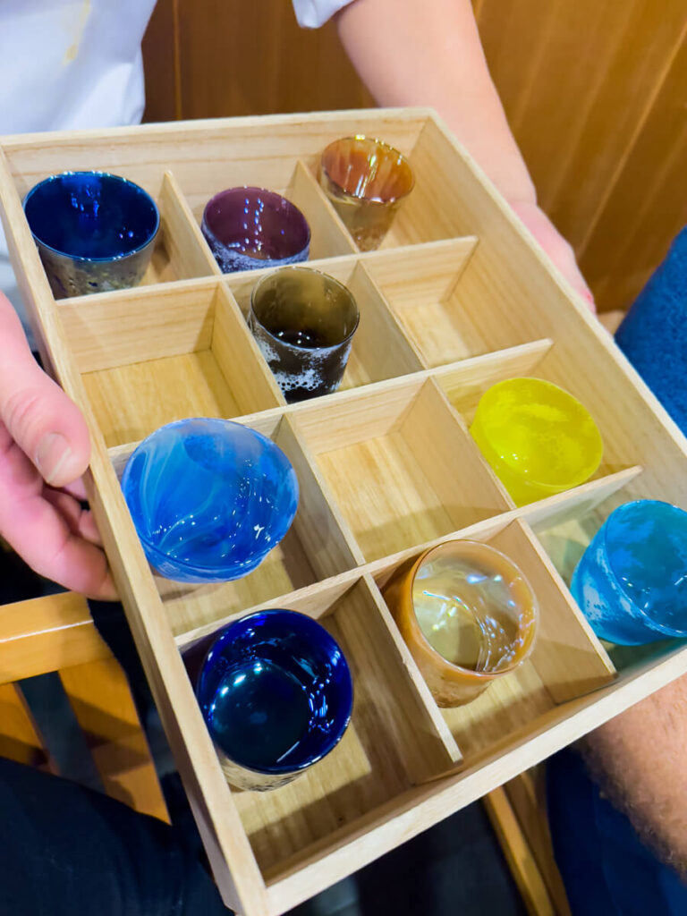 Colorful Japanese sake cups arranged in a wooden masu box with individual square compartments, each cup made of hand blown glass in shades of blue, amber, purple, and bright yellow. The box is held at a sushi counter, showcasing a curated sake tasting flight with vibrant glassware that highlights the craftsmanship and presentation of the dining experience.