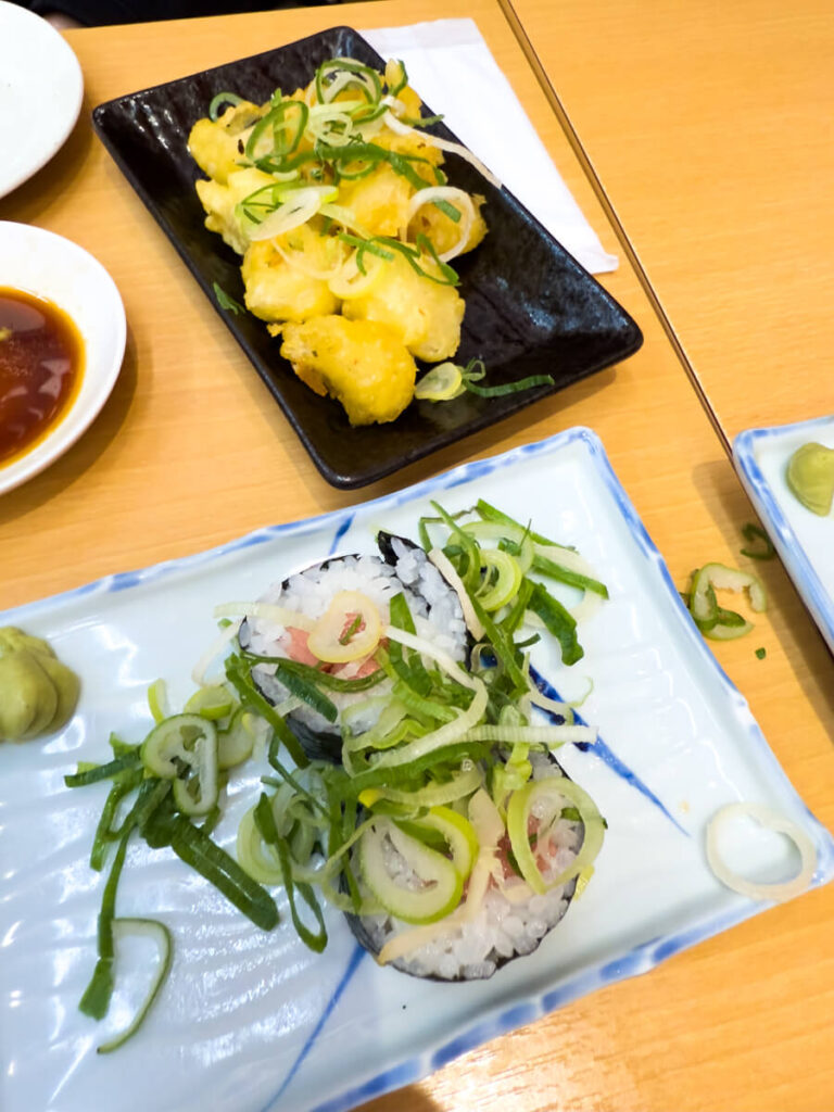 Black plate of sushi rolls topped with sliced green onions beside a dish of golden fried tempura scallops garnished with scallions. Small plates of soy sauce and wasabi sit nearby on the wooden table.