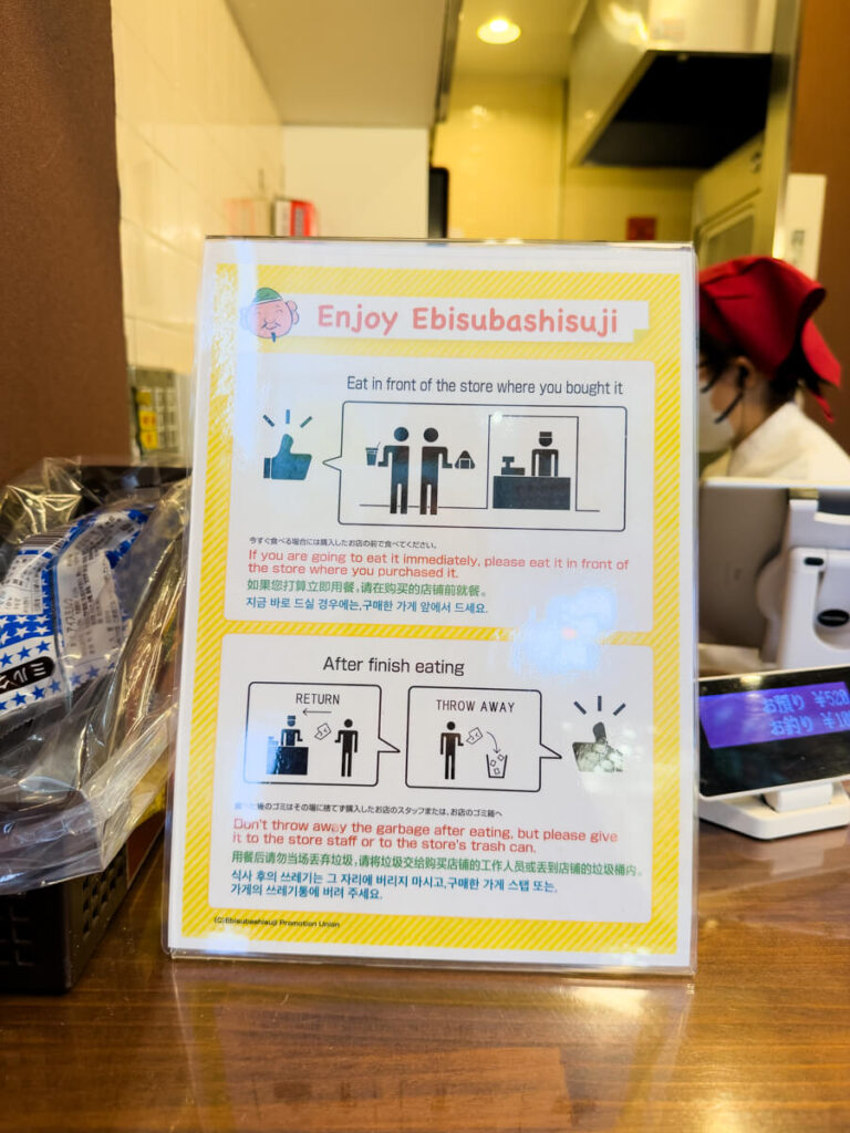 Sign at a food counter reading Enjoy Ebisubashisuji with instructions in English and other languages to eat in front of the store and return trash after finishing. The notice encourages proper etiquette for eating street food in the shopping area.