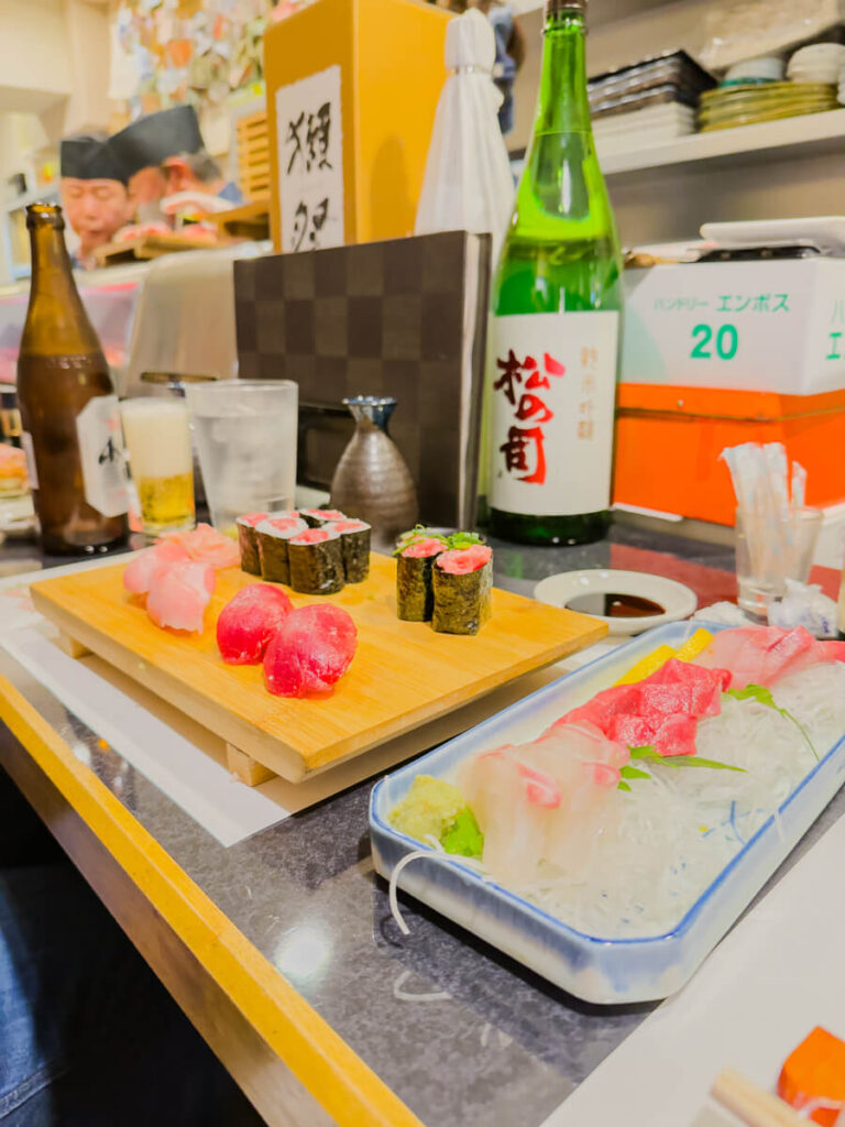 Sushi and sashimi spread at a counter with tuna nigiri maki rolls and sliced fish arranged on wooden boards. Bottles of sake beer and a small carafe sit behind the dishes creating a lively dining scene.
