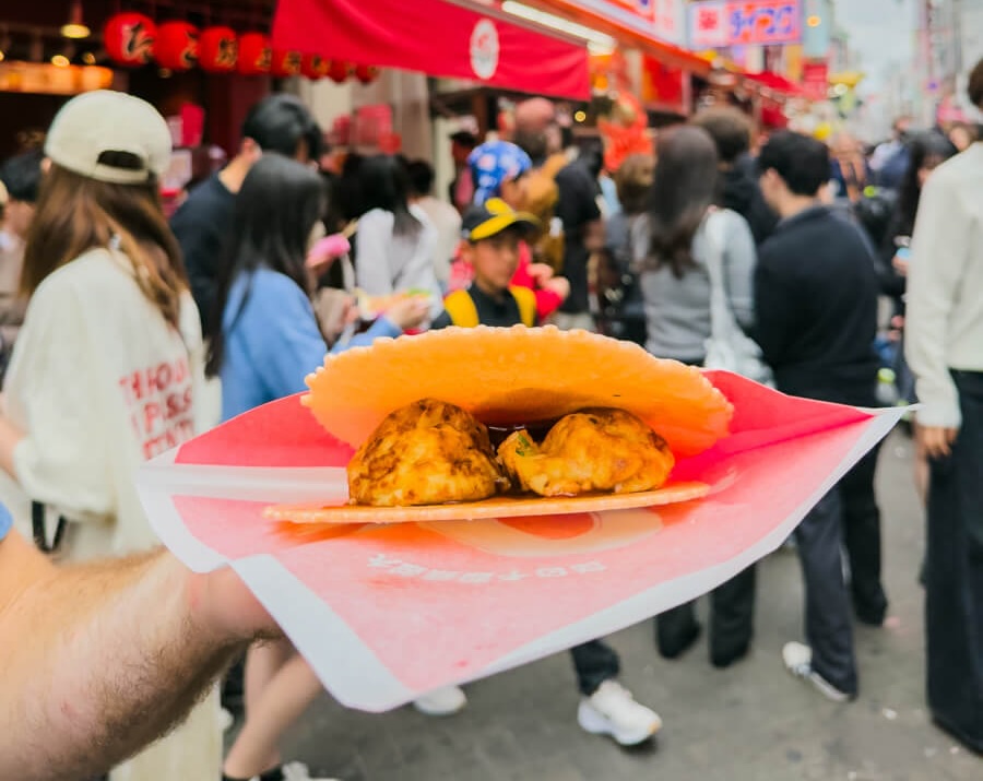 Takoyaki served in a paper tray on Dotonbori Street in Osaka Japan with two golden octopus balls topped in sauce and a large shrimp cracker resting on top. The street food is held up in the foreground while a busy crowd of shoppers and bright red storefront signs blur together in the background, capturing the lively atmosphere of this popular Osaka food district.