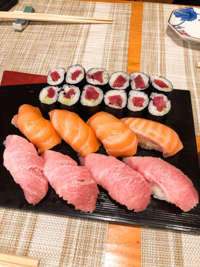 Assorted sushi platter featuring fatty tuna salmon nigiri and tuna rolls arranged neatly on a black rectangular board. The fresh fish sits over compact rice with chopsticks and small dishes set on the table.