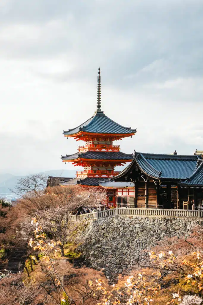 Iconic view of Kiyomizu-dera Temple in Kyoto with its red pagoda and traditional tiled rooftops surrounded by early cherry blossoms, a popular sight during sakura season in Japan.