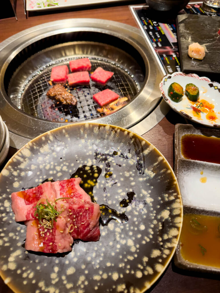 Thin slices of marbled beef cooking on a round tabletop grill surrounded by dipping sauces and small side dishes. The yakiniku setup shows the meat sizzling over open flame at the center of the table.