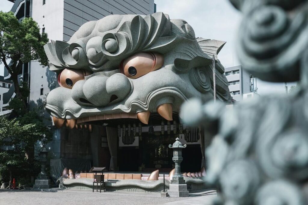 Namba Yasaka Shrine in Osaka featuring the massive lion head stage with wide open mouth, large round eyes, and prominent fangs forming the entrance to the shrine hall. The dramatic Shinto shrine architecture stands in a quiet courtyard surrounded by city buildings, highlighting the unique and photogenic design of Namba Yasaka Shrine.
