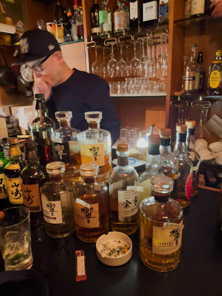 Cozy bar interior with numerous bottles of Japanese whisky arranged on the counter, including Hibiki and other labels. A bartender wearing a cap stands behind the bar with hanging glassware and shelves of liquor bottles in the background.