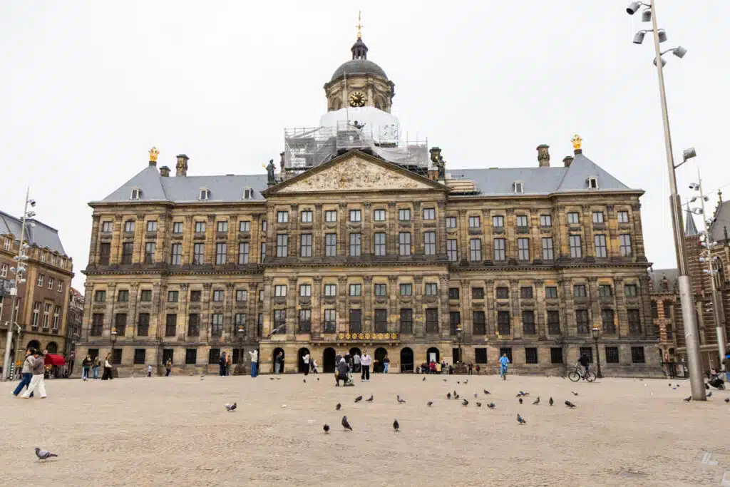 The Royal Palace on Dam Square in central Amsterdam with its grand stone facade and clock tower rising above an open plaza where people walk and pigeons gather. This landmark is a key stop on any Amsterdam itinerary and sits at the heart of the city’s historic center.