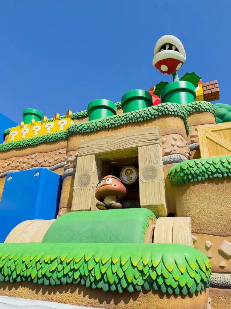 Goomba character standing at the entrance of a small tunnel in Super Nintendo World at Universal Studios Japan in Osaka surrounded by green hills question blocks pipes and a Piranha Plant above.