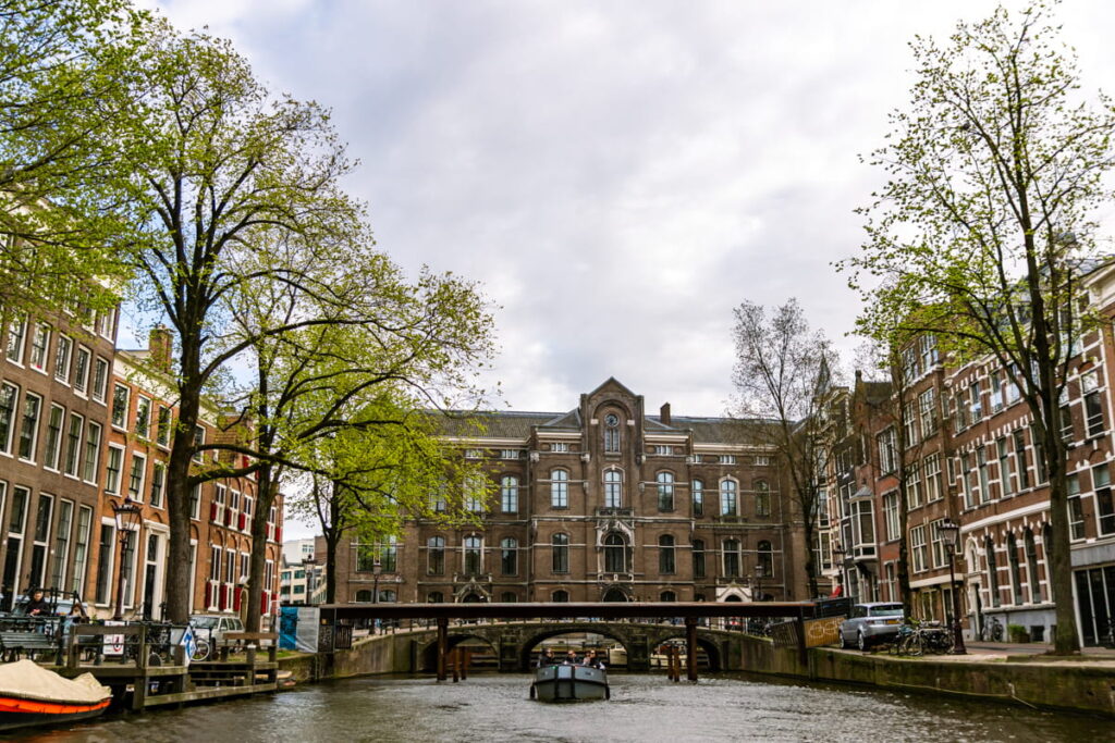 boat crossing under a bridge in Amsterdam with large green trees on either side and a building in the background