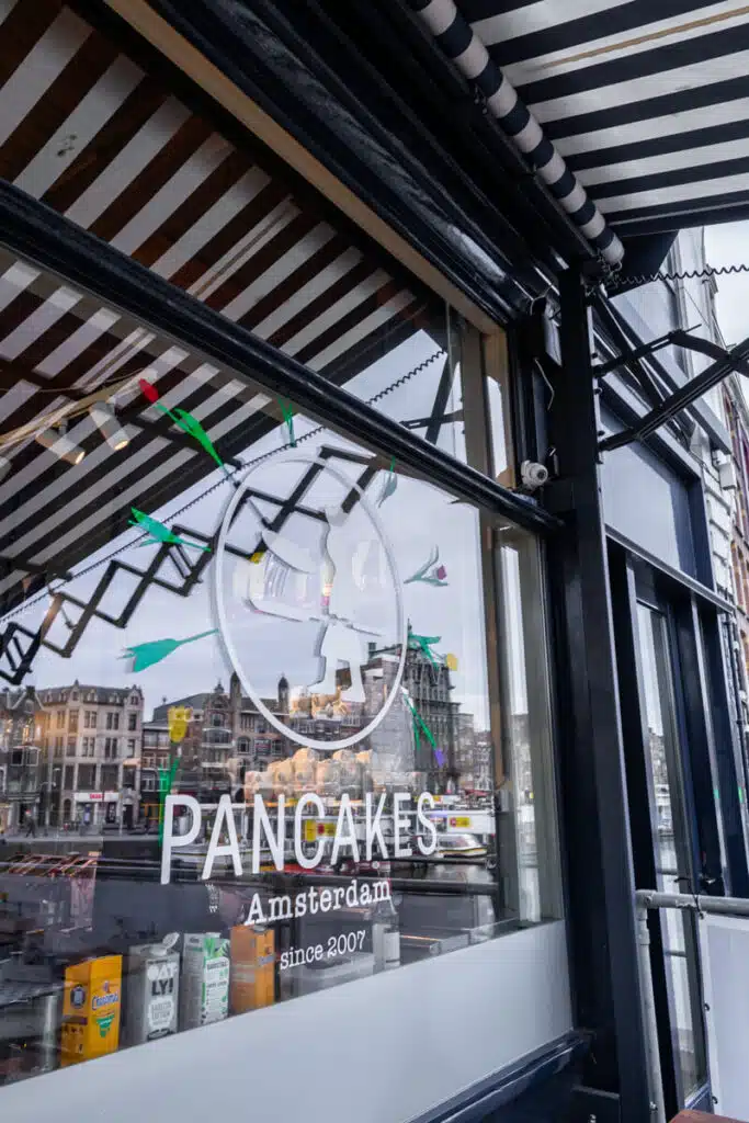 Window of Pancakes Amsterdam with striped awning and reflections of canal houses across the water with text that reads "Pancakes Amsterdam" and "Since 2007". This spot is a popular food stop to include on an Amsterdam itinerary.