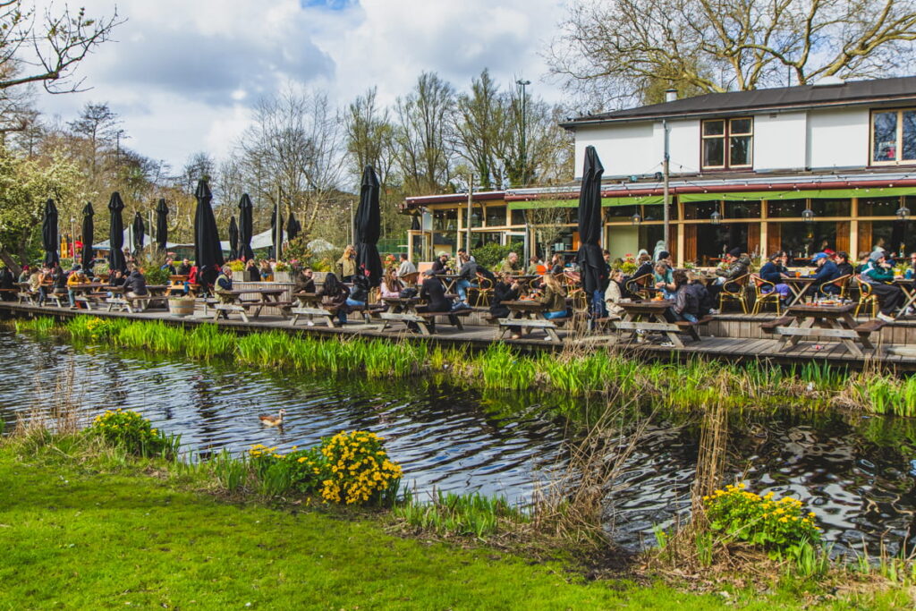 Outdoor terrace at Vondelpark beside a canal with people sitting at picnic tables under black umbrellas enjoying food and drinks. A duck swims in the water in front of the busy Groot Melkhuis café scene on a sunny day in Amsterdam.