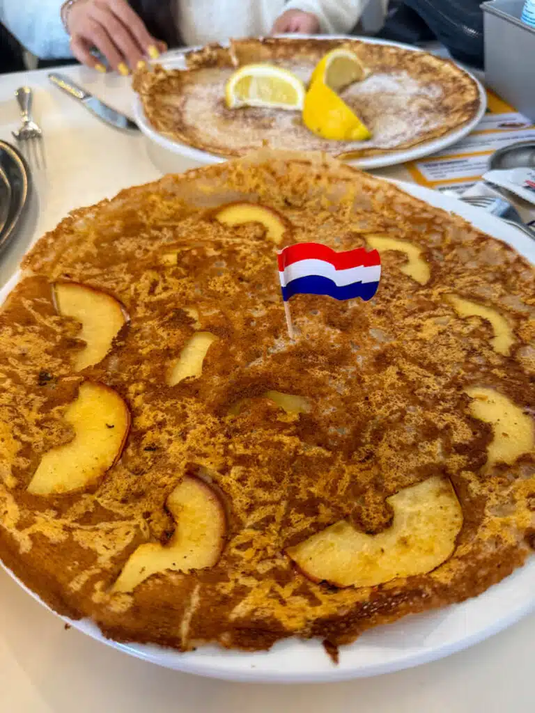Large Dutch apple pancake topped with cinnamon and sliced apples served on a white plate with a small Dutch flag and a second pancake with lemon slices in the background. Trying traditional pancakes like this is a delicious experience to have in Amsterdam