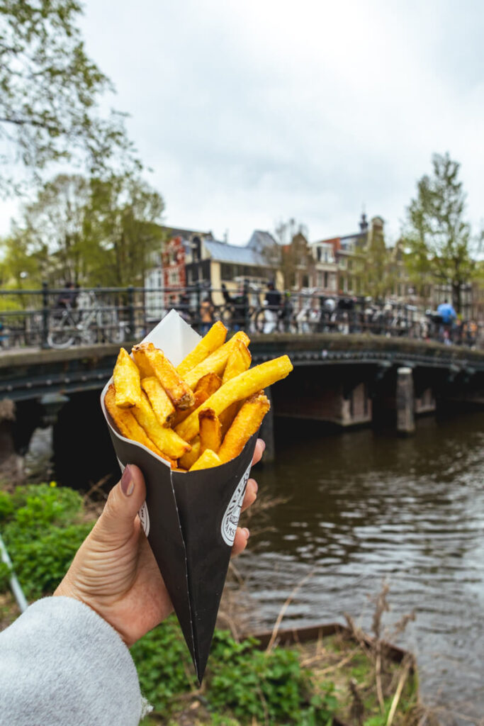 Hand holding a cone of golden fries from Heertje Friet near a canal bridge in Amsterdam with bikes and water in the background. Grabbing fries by the canal is a classic casual bite on an Amsterdam itinerary.