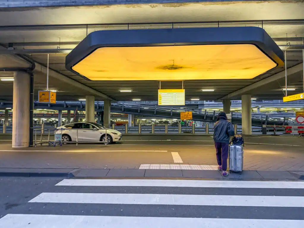 Traveler pulling a suitcase across a pedestrian crossing at Amsterdam Airport Uber pick up area under a large glowing yellow ceiling light with parking signs and cars in the background. This scene shows the transition from airport to city at the start of an Amsterdam itinerary.