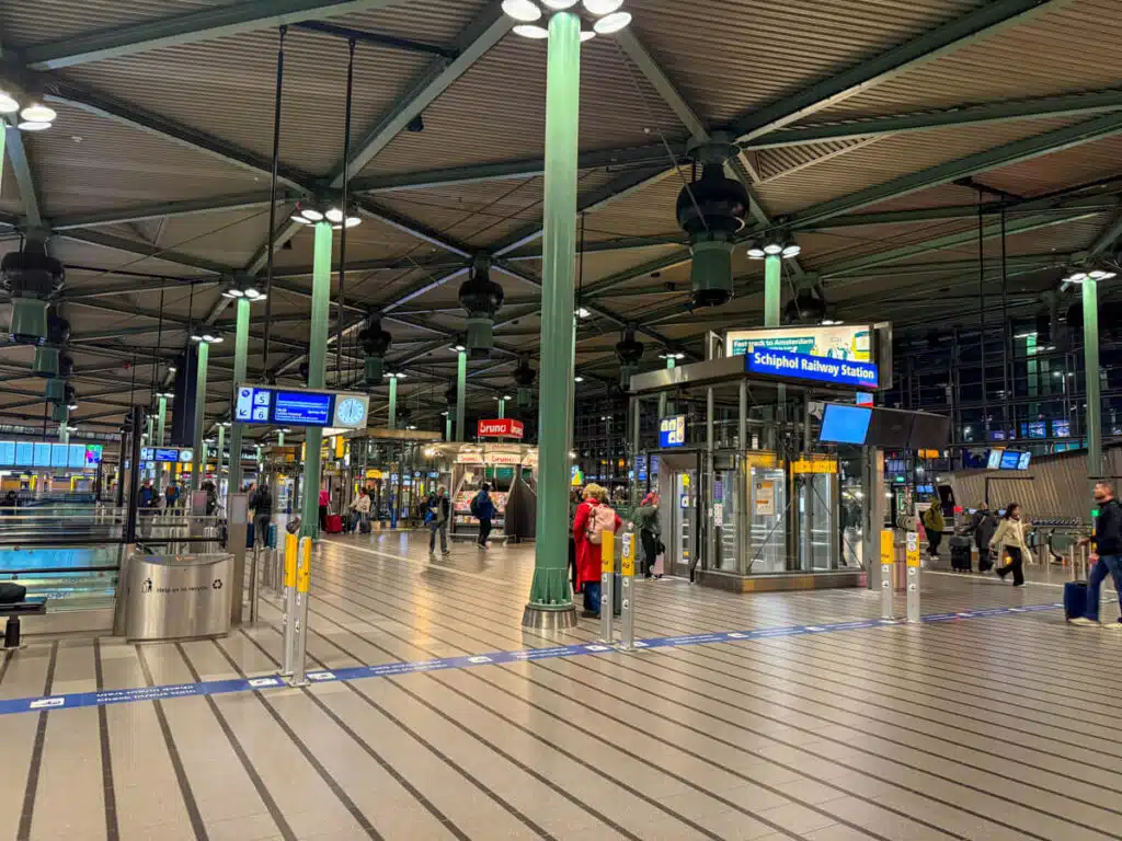 Interior of Schiphol Railway Station at Amsterdam Airport with green steel columns striped gray flooring and a few travelers walking between platforms and ticket gates. Blue signs read "Schiphol Railway Station" and this transit hub is a common first stop on an Amsterdam itinerary.
