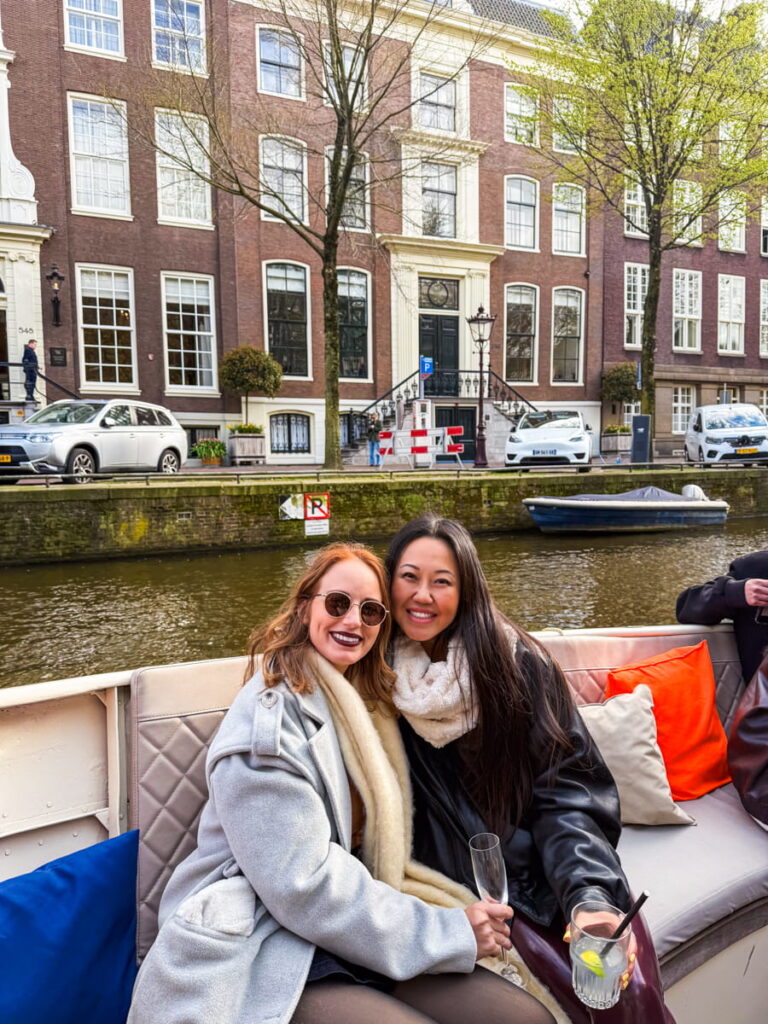 Two women sit together on a canal cruise boat in Amsterdam smiling while holding drinks with canal houses and parked cars behind them. Cushioned seating and colorful pillows create a relaxed atmosphere as they enjoy the ride.