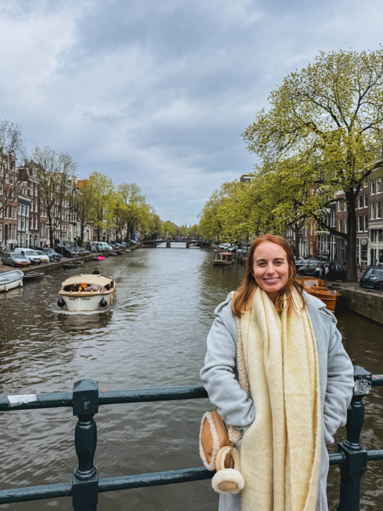 Woman in a coat and scarf smiling on a canal bridge in Amsterdam with boats passing behind her and rows of houses lining the water. This captures a typical moment while exploring Jordaan on an Amsterdam itinerary.