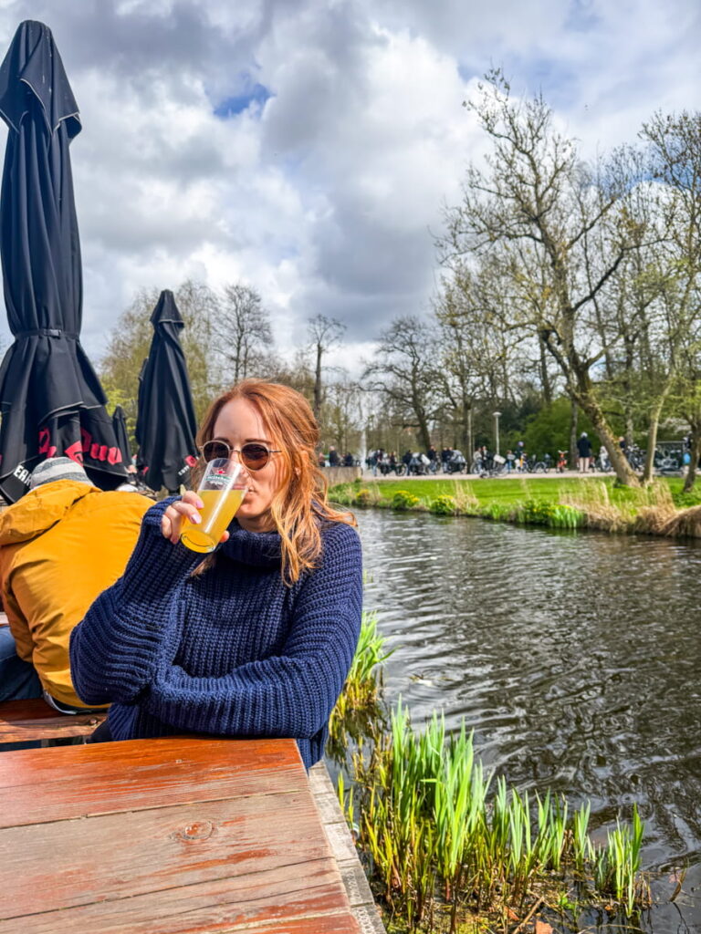 Woman wearing sunglasses and a navy sweater drinks a beer at a wooden table beside the water at Groot Melkhuis café in Vondelpark. The canal and park with cyclists and greenery are visible behind her on a bright day in Amsterdam.