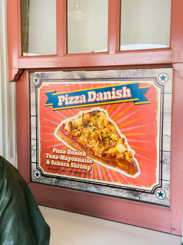 Food sign at Universal Studios Japan in Osaka reading "Pizza Danish Tuna Mayonnaise & Sakura Shrimp" with an image of a slice of pastry topped with seafood and herbs.