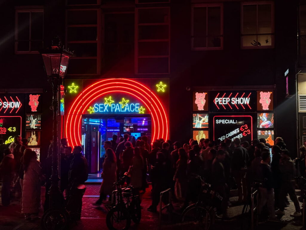 Crowded street in Amsterdam’s Red Light District at night with neon signs reading "Sex Palace" and "Peep Show" illuminating the scene in red and blue lights. Exploring this lively area is a well known experience on an Amsterdam itinerary.