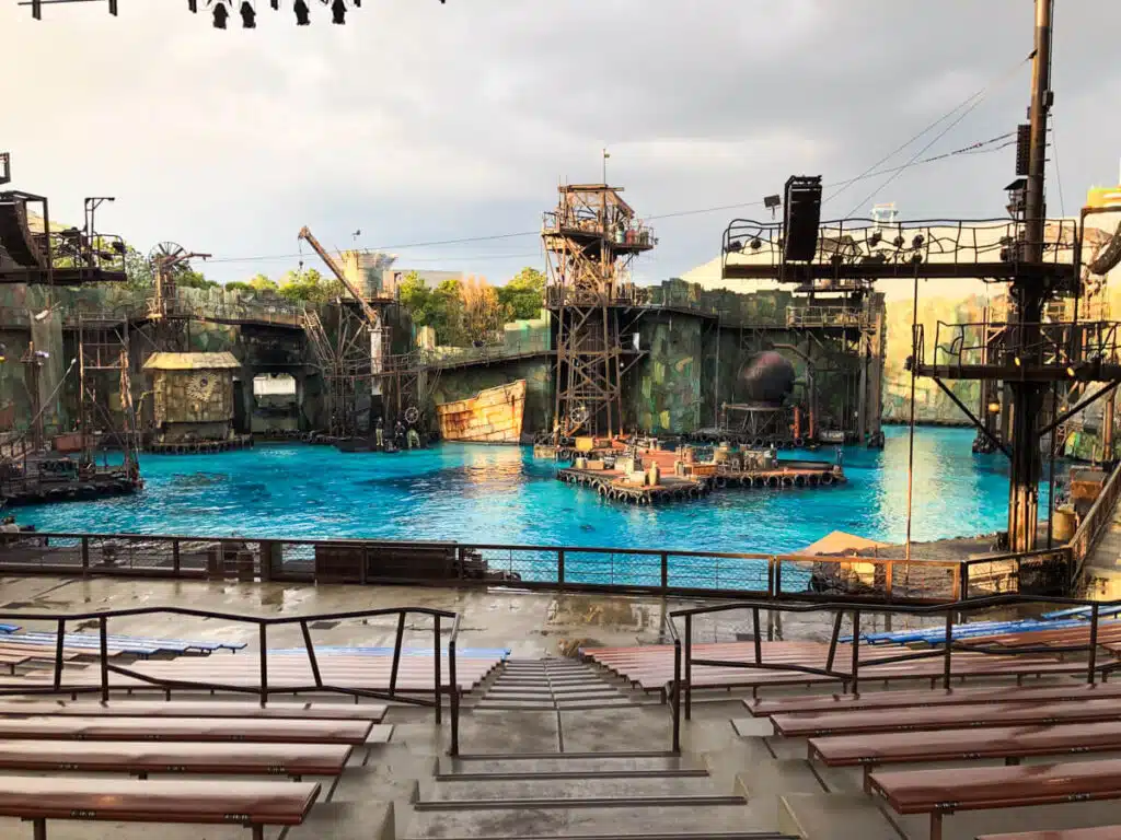 WaterWorld stunt show arena at Universal Studios Japan in Osaka with a large pool industrial set structures and empty stadium seating in the foreground before the performance begins.