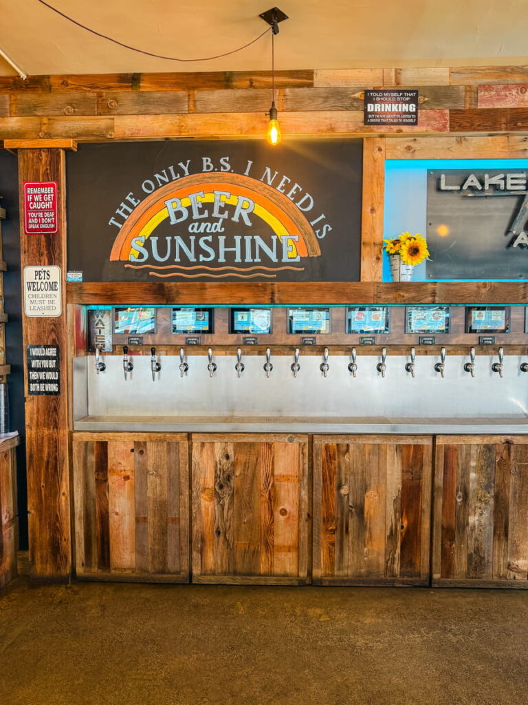 Self serve tap wall at Lake Tahoe AleWorX with wooden panels and a sign reading "THE ONLY B.S. I NEED IS BEER and SUNSHINE" above rows of beer taps and digital screens.