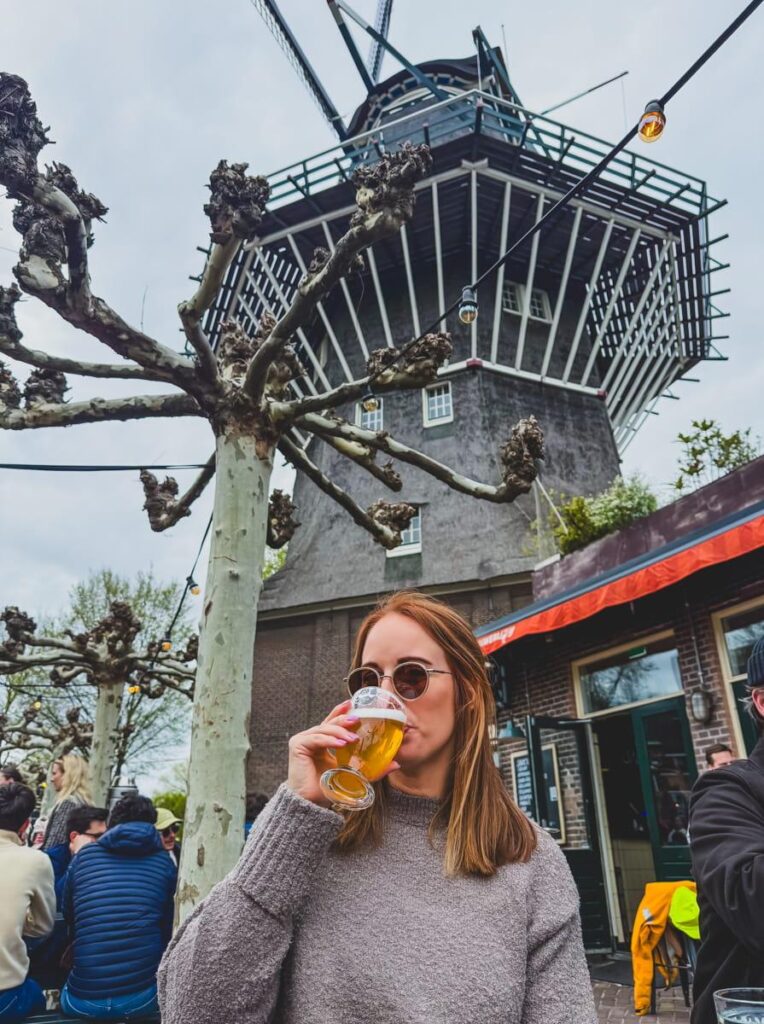 Woman in sunglasses sipping a glass of beer while seated outside Brouwerij 't IJ windmill brewery with the iconic windmill towering behind her.