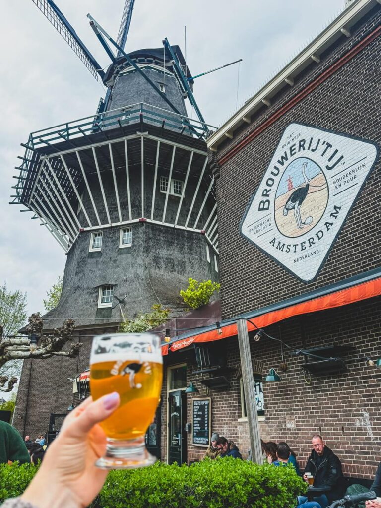 Hand holding a glass of beer in front of Brouwerij 't IJ windmill brewery with the large traditional windmill and brewery building in the background. This view captures one of the most unique brewery settings in Amsterdam.