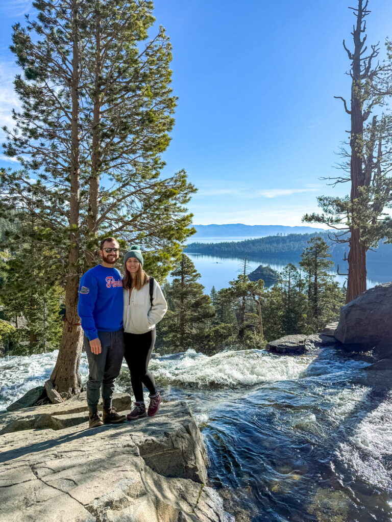couple stands next to the top of Eagle Falls with trees and Emerald Bay in the background