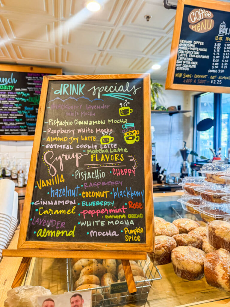 Chalkboard at Tahoe Bagel Company with colorful handwritten "drink specials" and "syrup flavors" displayed above a counter of fresh baked goods