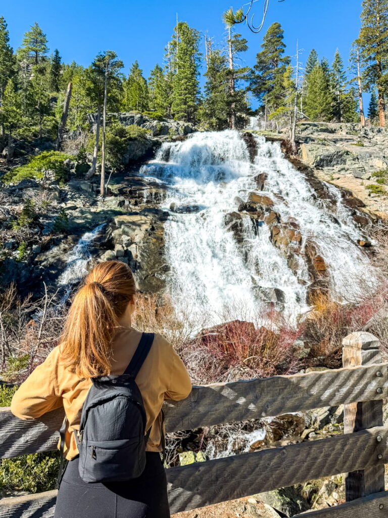 girl in yellow shirt with black backpack stands on a wooden bridge in front of Eagle Falls in Lake Tahoe