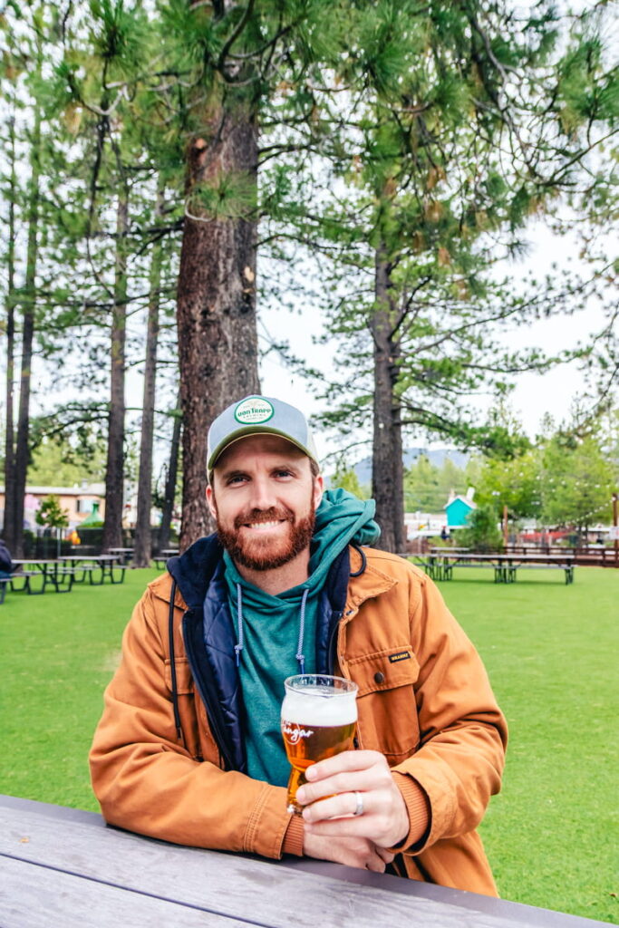 men sitting at picnic table at the Hangar Taproom in Lake Tahoe holding a beer with large grassy green space and trees behind him