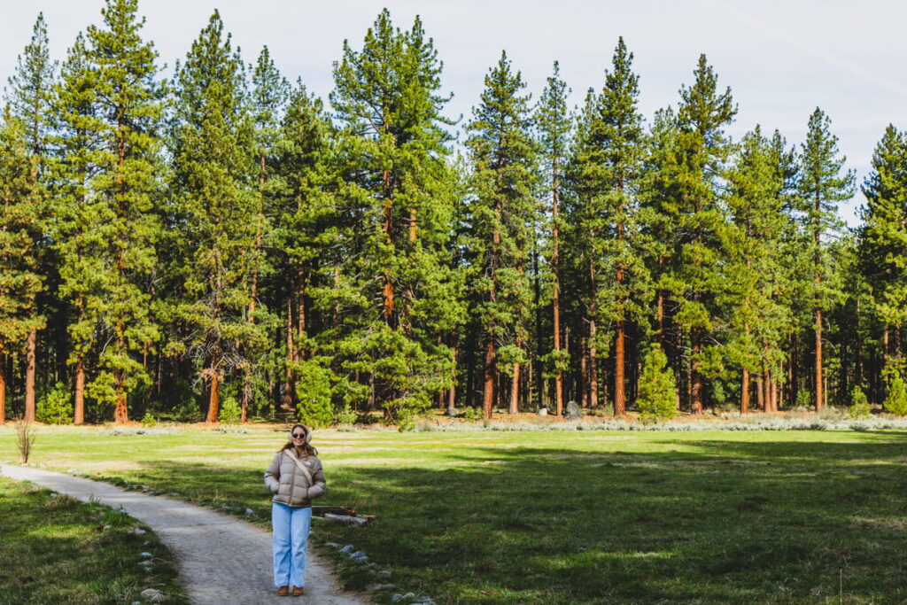 girl standing on a path in large meadow with large pine trees in background along the Lam Watah Trail